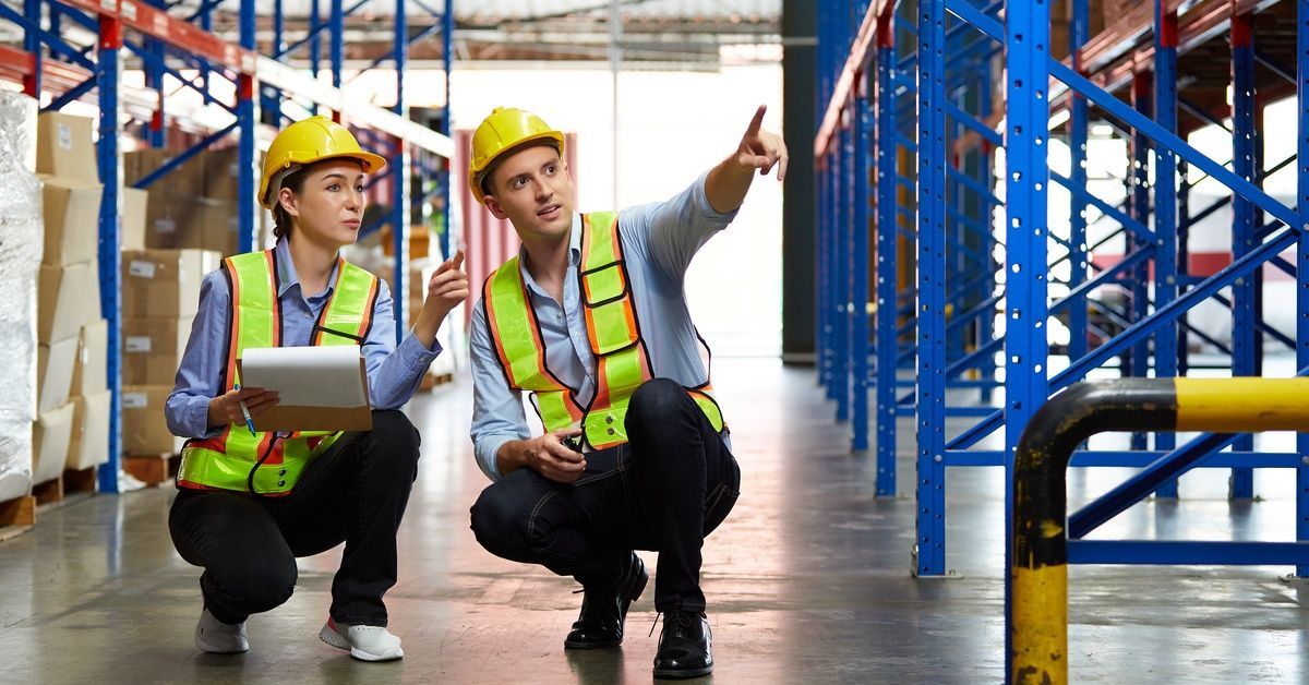 Two factory workers crouched to look at different parts of the pallet rack. Both employees are wearing safety gear.