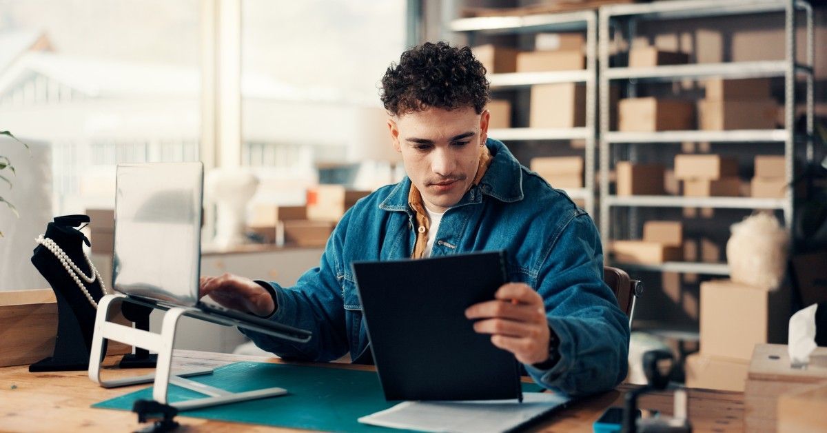 A man working at a desk with shelving units behind him. He is looking at a paper while typing on a laptop.