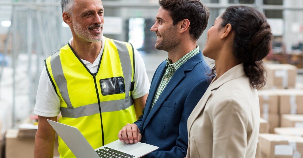 A worker and two managers are discussing something in a warehouse. One of the managers is holding a laptop.