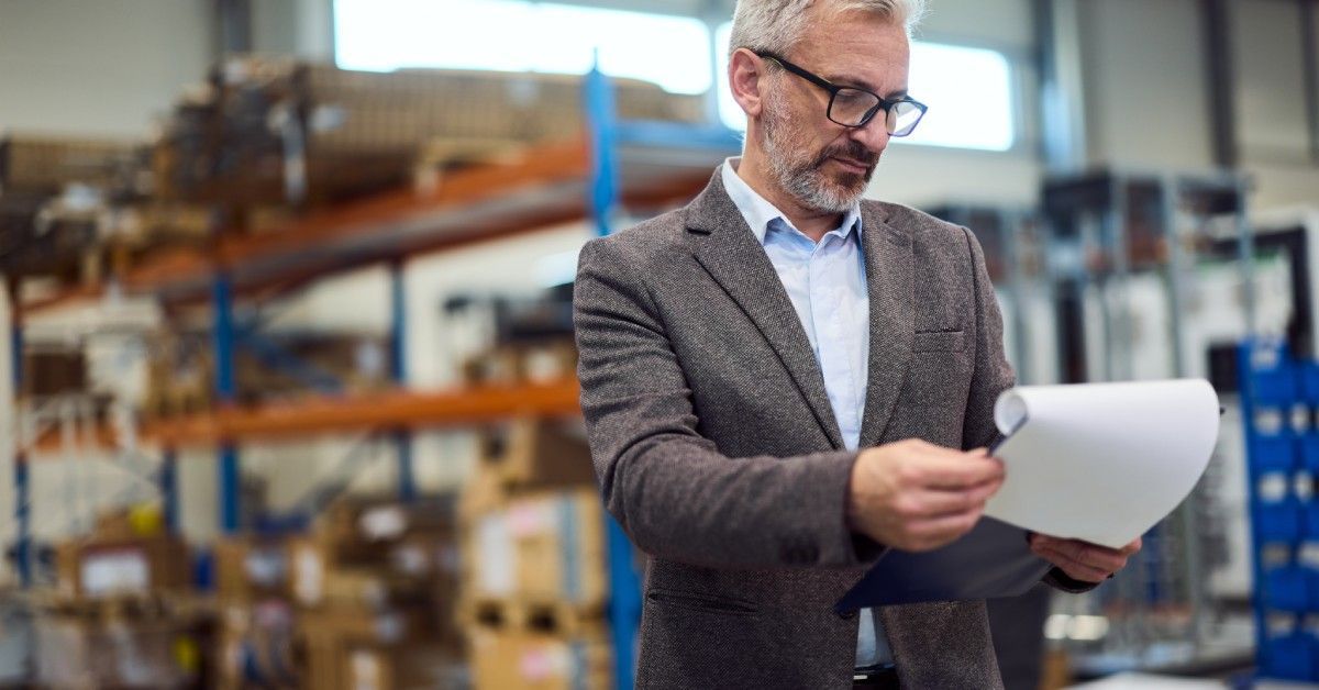 A business professional in a warehouse setting. He is looking at a paper on a clipboard.