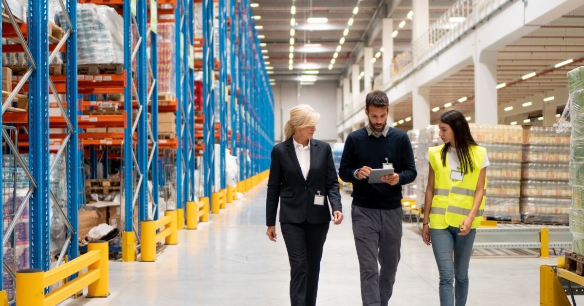 Two managers walk around a warehouse with an employee. The managers are wearing business casual outfits.