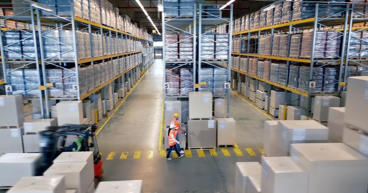 A bustling warehouse contains tall shelves stacked with inventory. Some employees are walking past the shelving units.