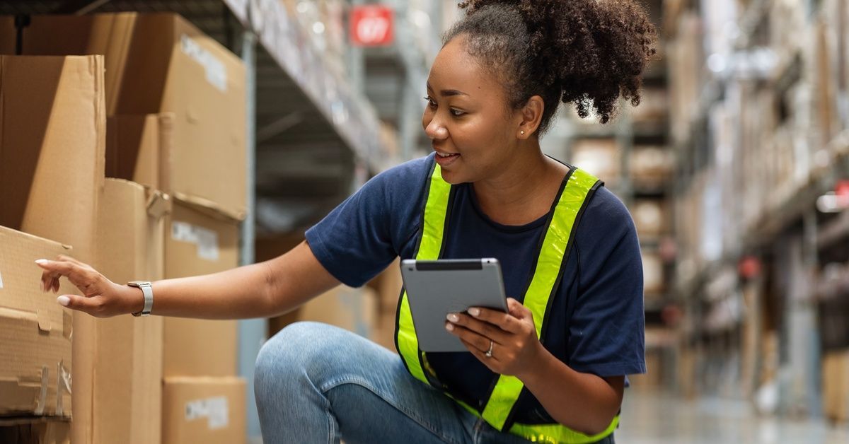 A young woman holding a tablet and checking the inventory on a warehouse's shelf. She is wearing a neon safety vest.