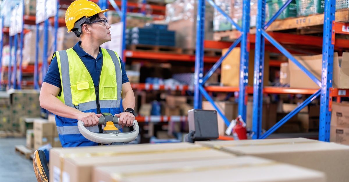 A worker moving a large cart of boxes through a warehouse. The worker is wearing a safety vest and a
