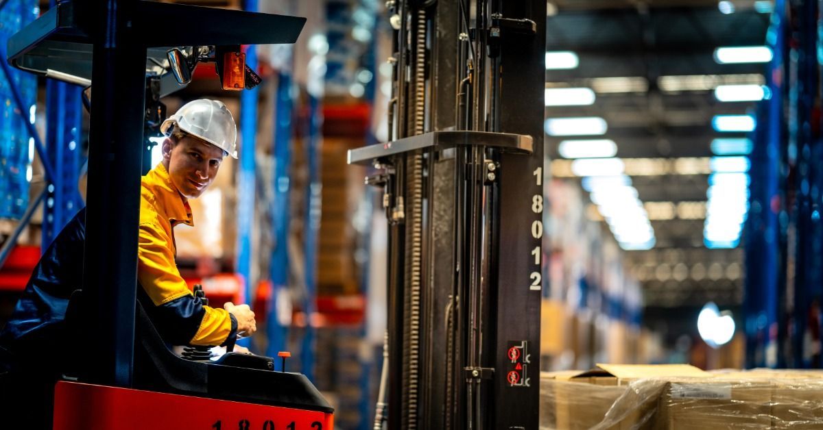 An industrial forklift driver pushing large boxes through a warehouse with tall shelving units behind it.