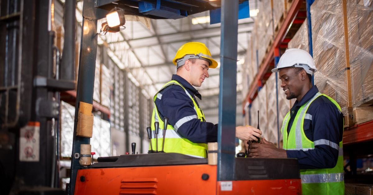 Two warehouse workers, with one of them standing in a forklift, move large packages around the facility.
