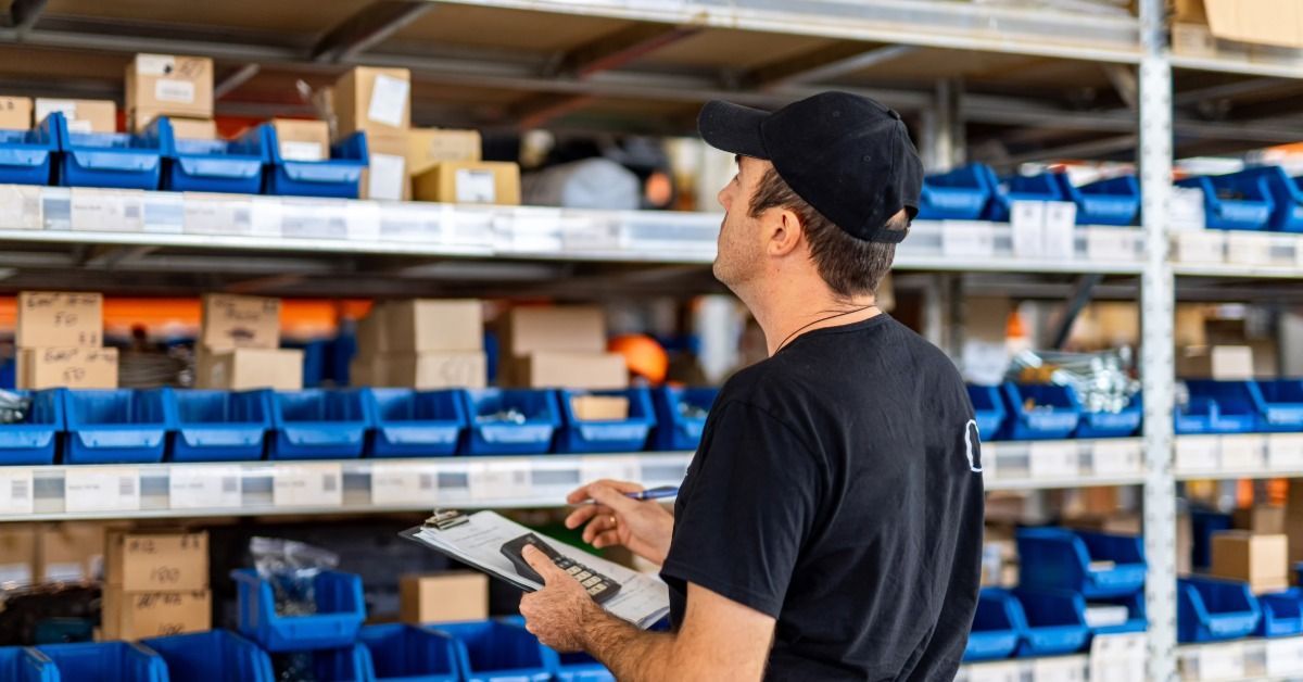 A warehouse worker standing in front of shelves with small bins. The worker is checking the inventory with a clipboard.