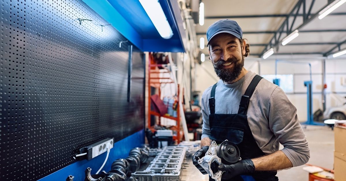 A happy car mechanic holding a piece of equipment. He is next to a workbench in an auto repair shop.