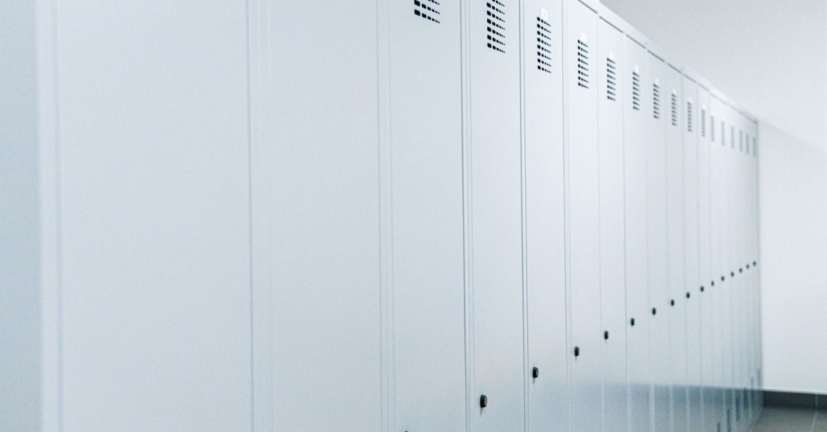 A set of white metal lockers off to the side of a wall in an office building. The lockers are all shut and locked.