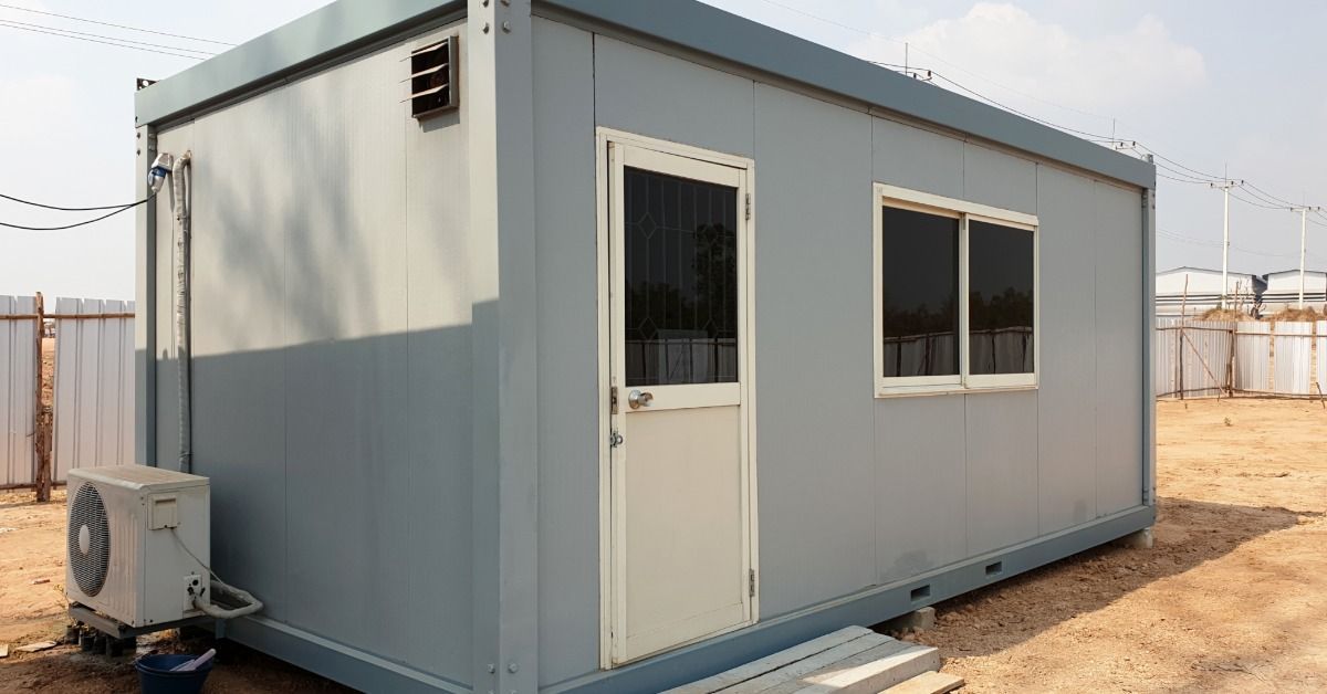 A gray rectangular modular office sits outside in the sand.
