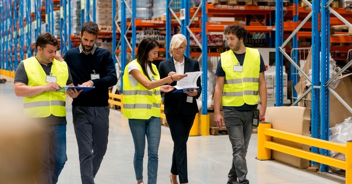 Workers in a warehouse walking with managers. They are looking through paperwork as they walk past t