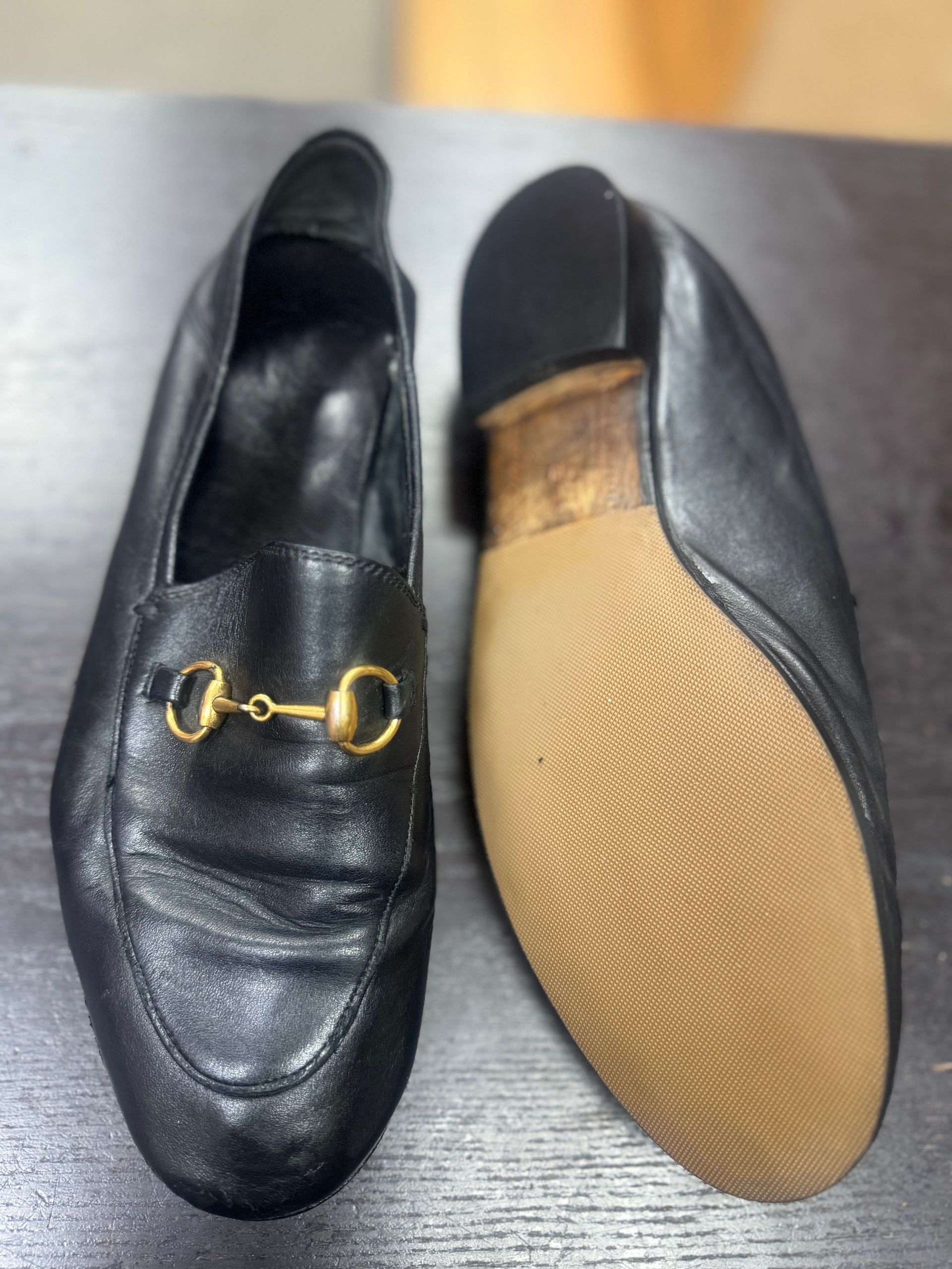 A pair of black leather loafers with gold horsebit detailing, one showing the upper and the other showing the sole.