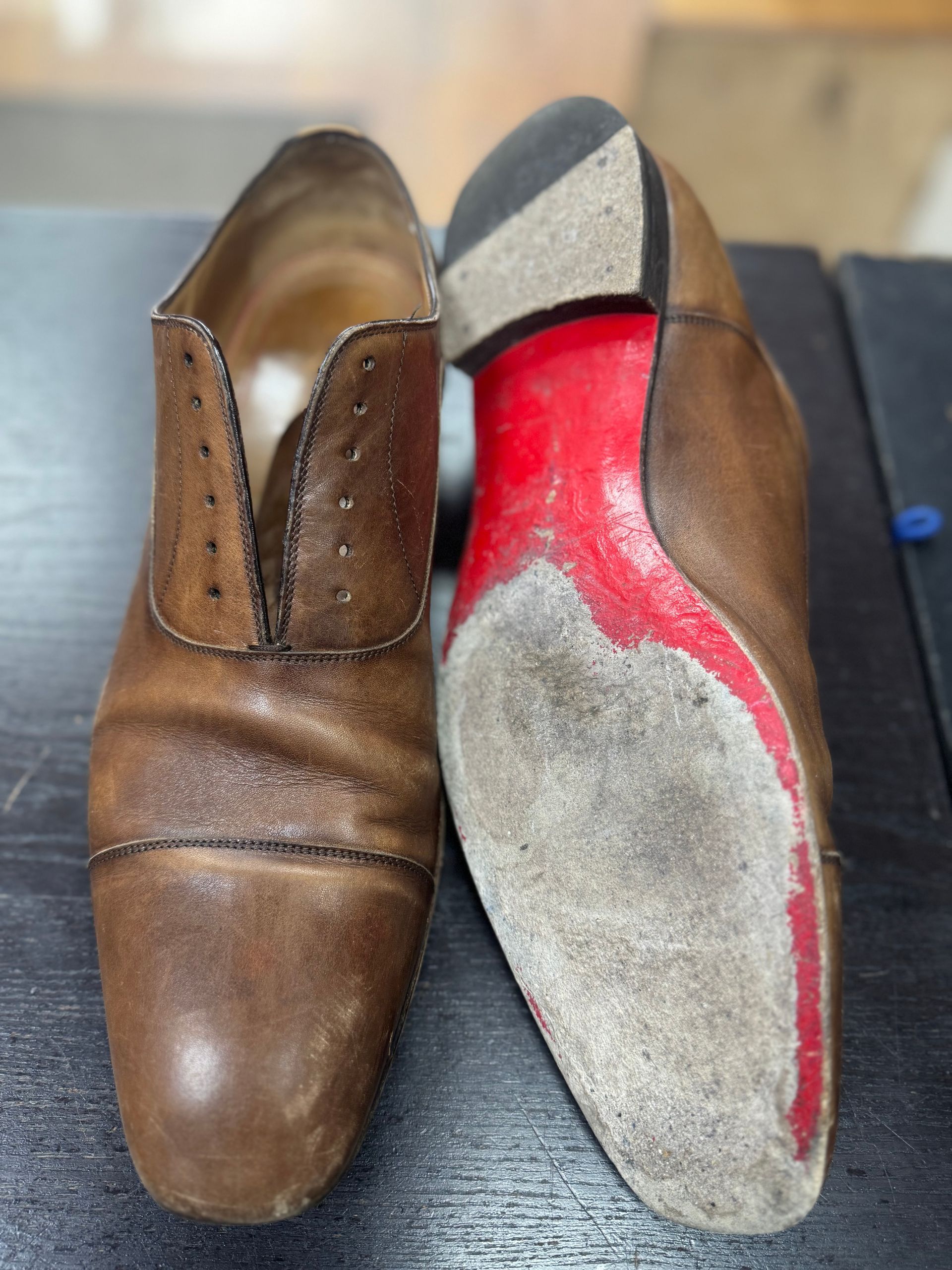 A pair of tan leather dress shoes, one showing the toe and the other flipped to reveal a worn red-painted sole.