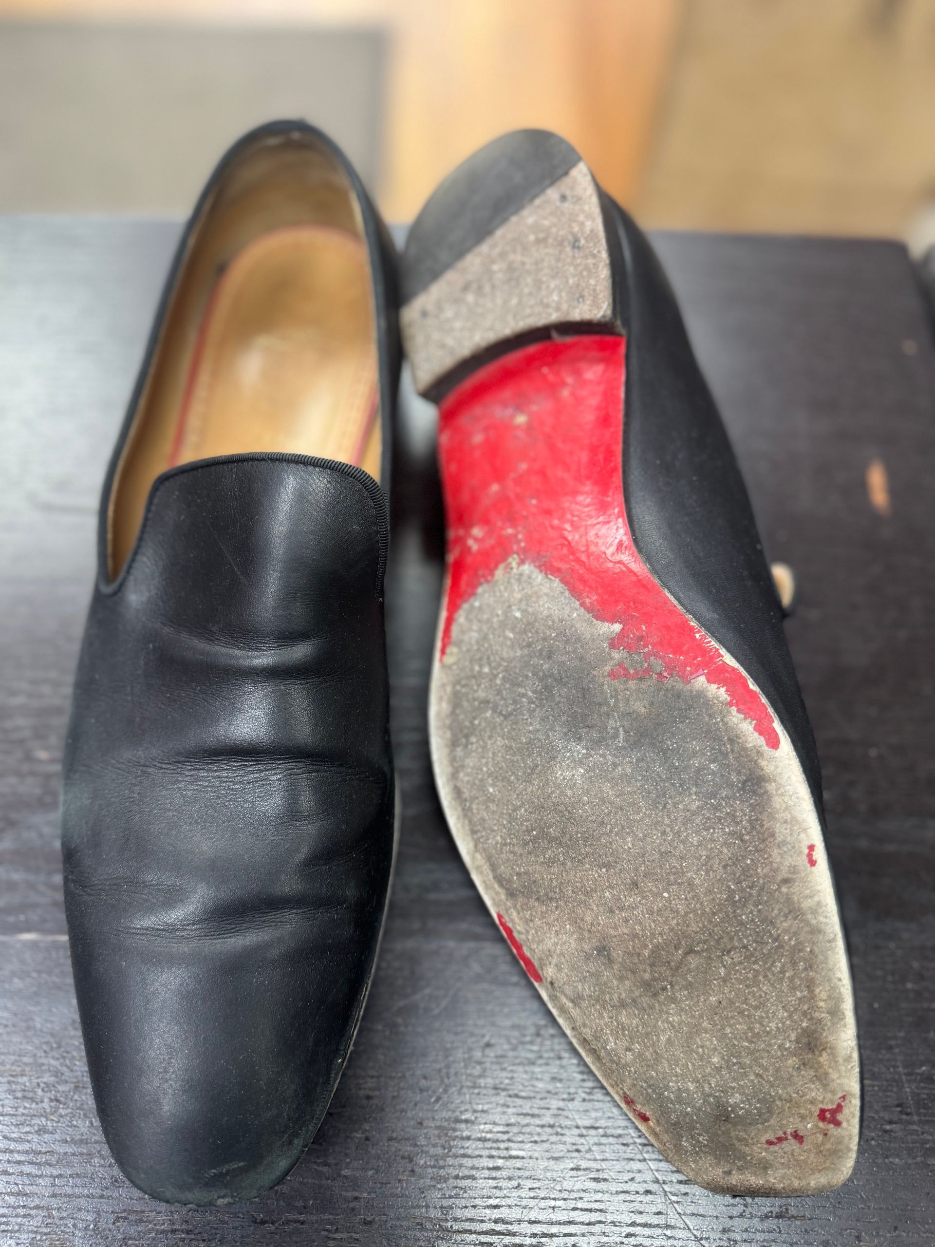 A pair of black leather loafers resting on a dark surface, with one shoe showing its worn red-painted sole.