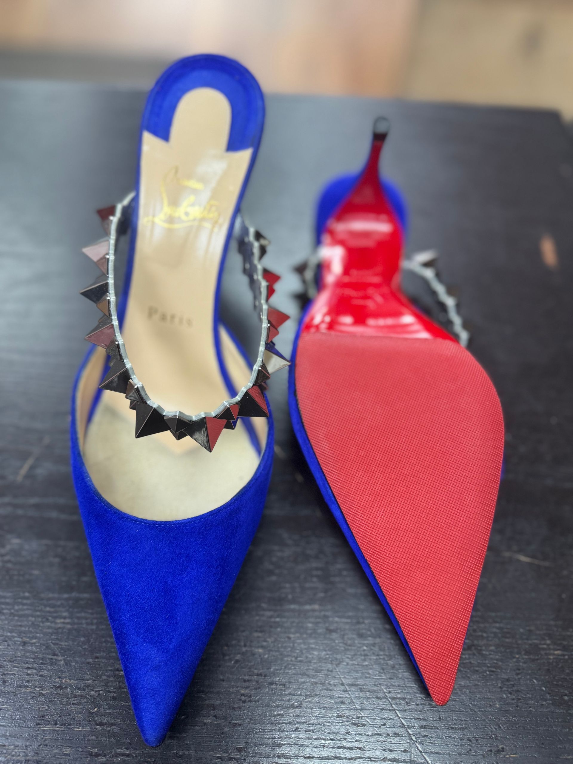 A pair of bright blue suede stiletto mules with black spike-studded straps and signature red soles on a dark surface.