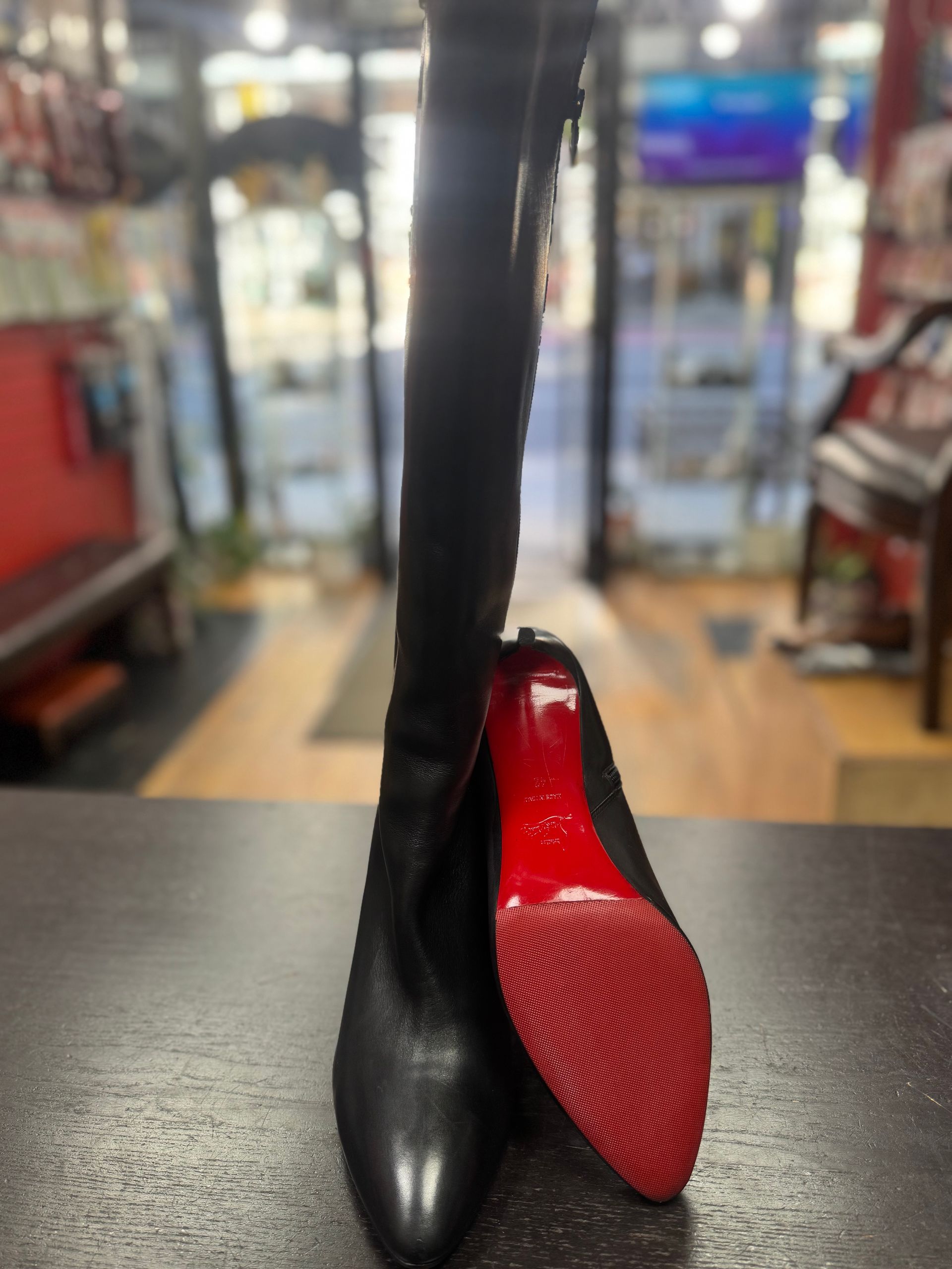 A black leather knee-high boot standing upright next to its detached, red-soled heel on a dark table in a shop.