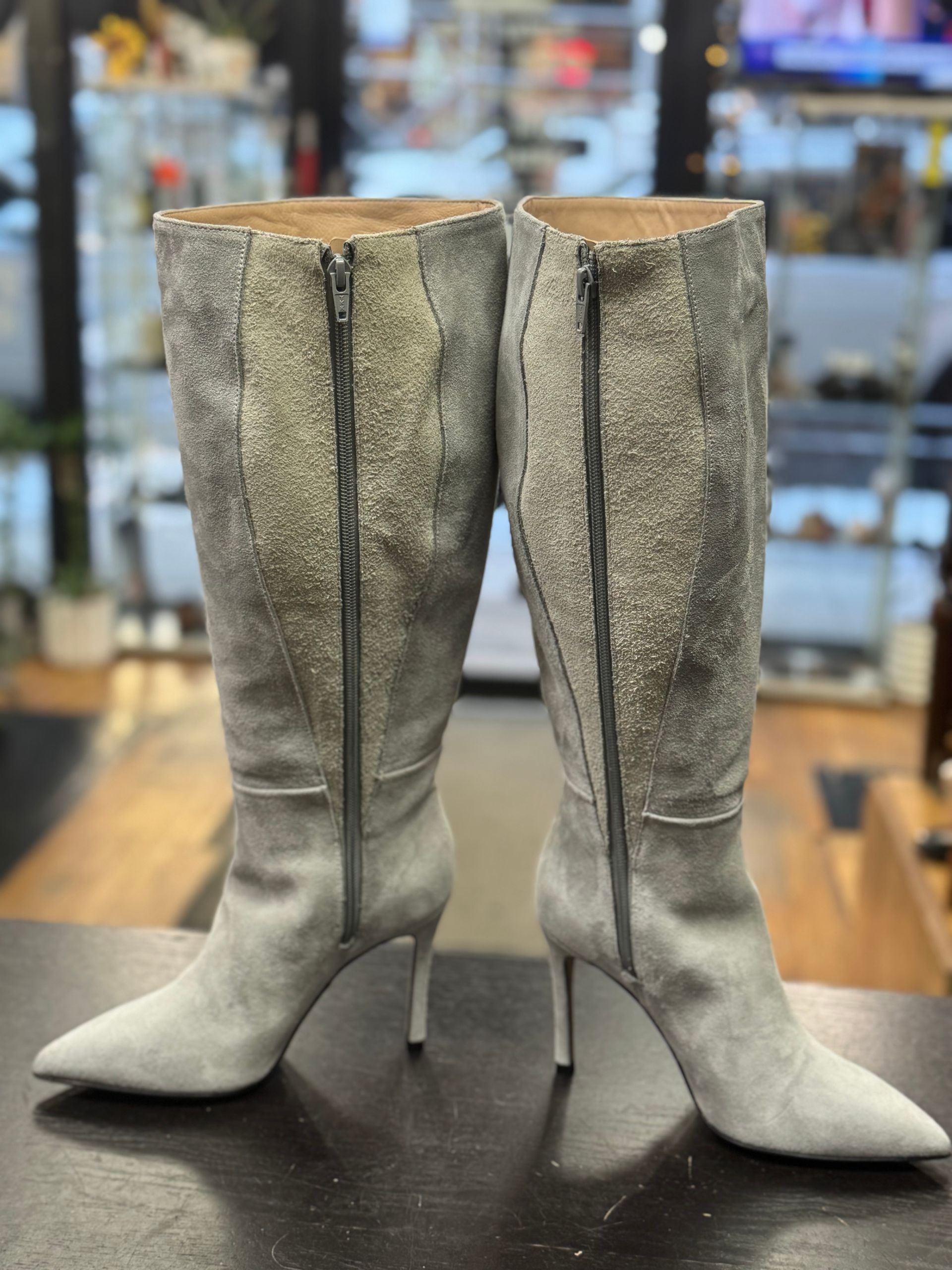 A pair of light gray suede, knee-high boots with thin high heels and full-length side zippers, displayed on a table.
