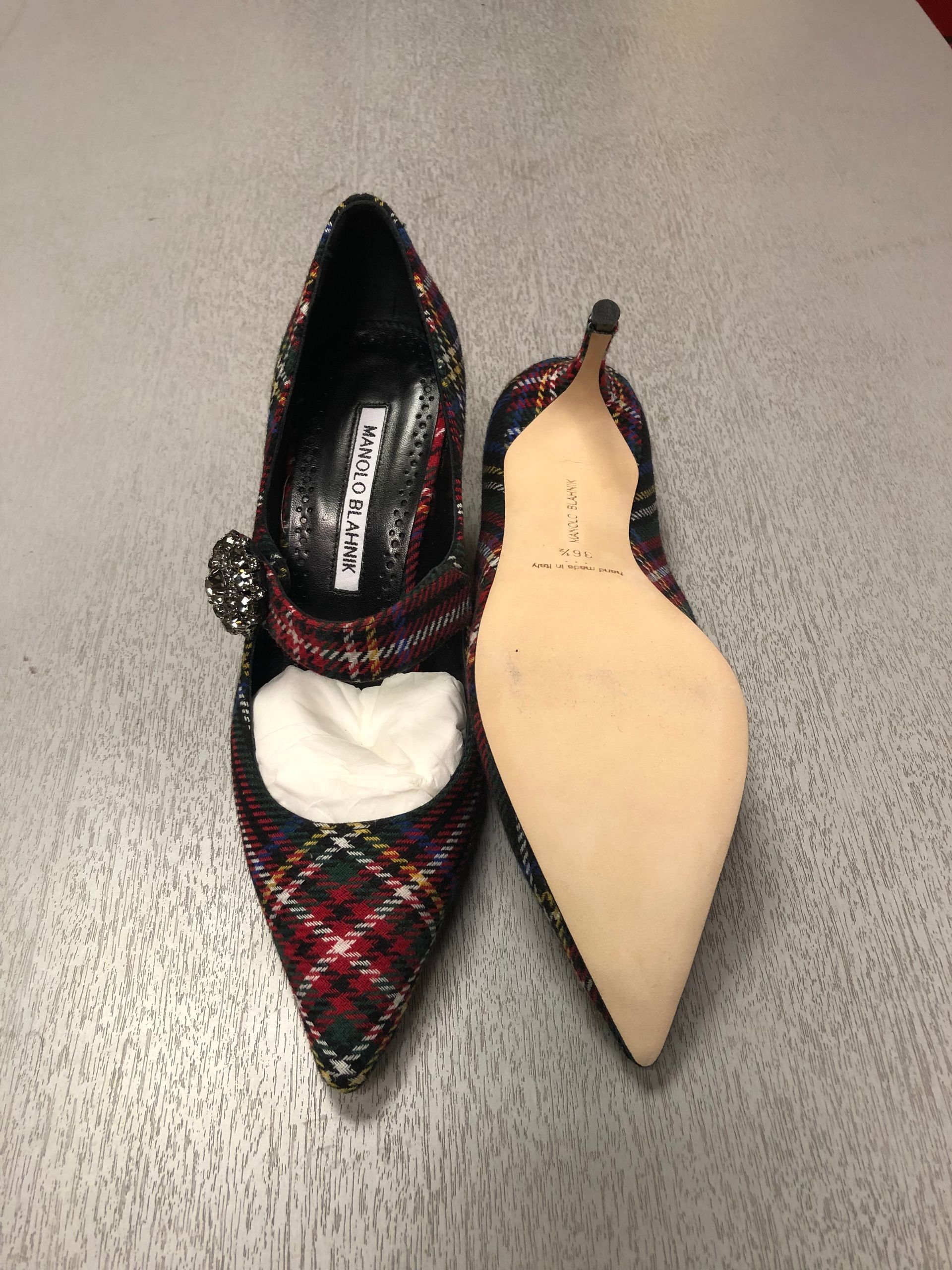 A pair of plaid Manolo Blahnik pointed-toe pumps with a crystal embellishment on a gray surface.