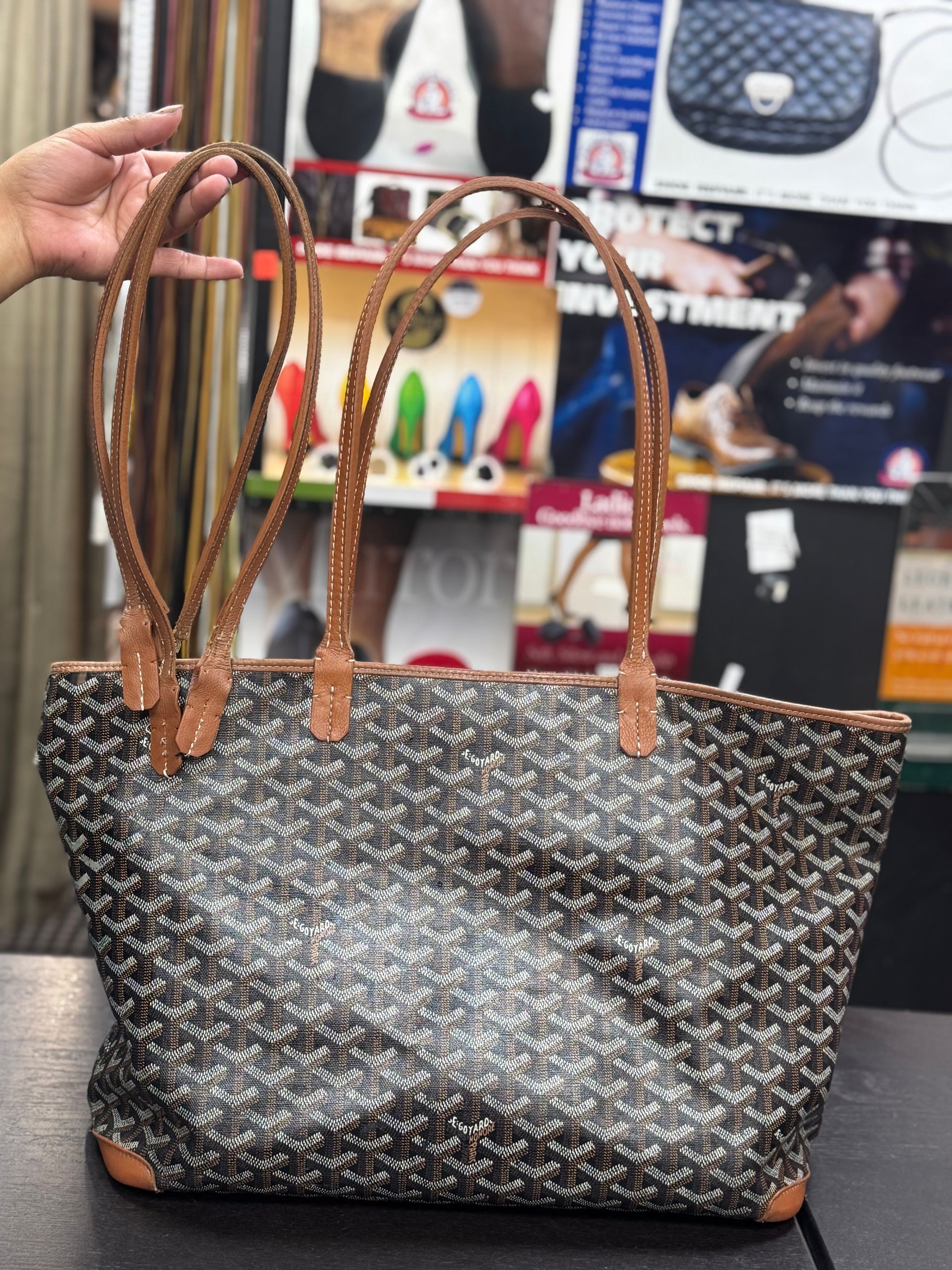 A hand holds a gray and black Goyard tote bag with brown leather handles and base, displayed in a retail store.