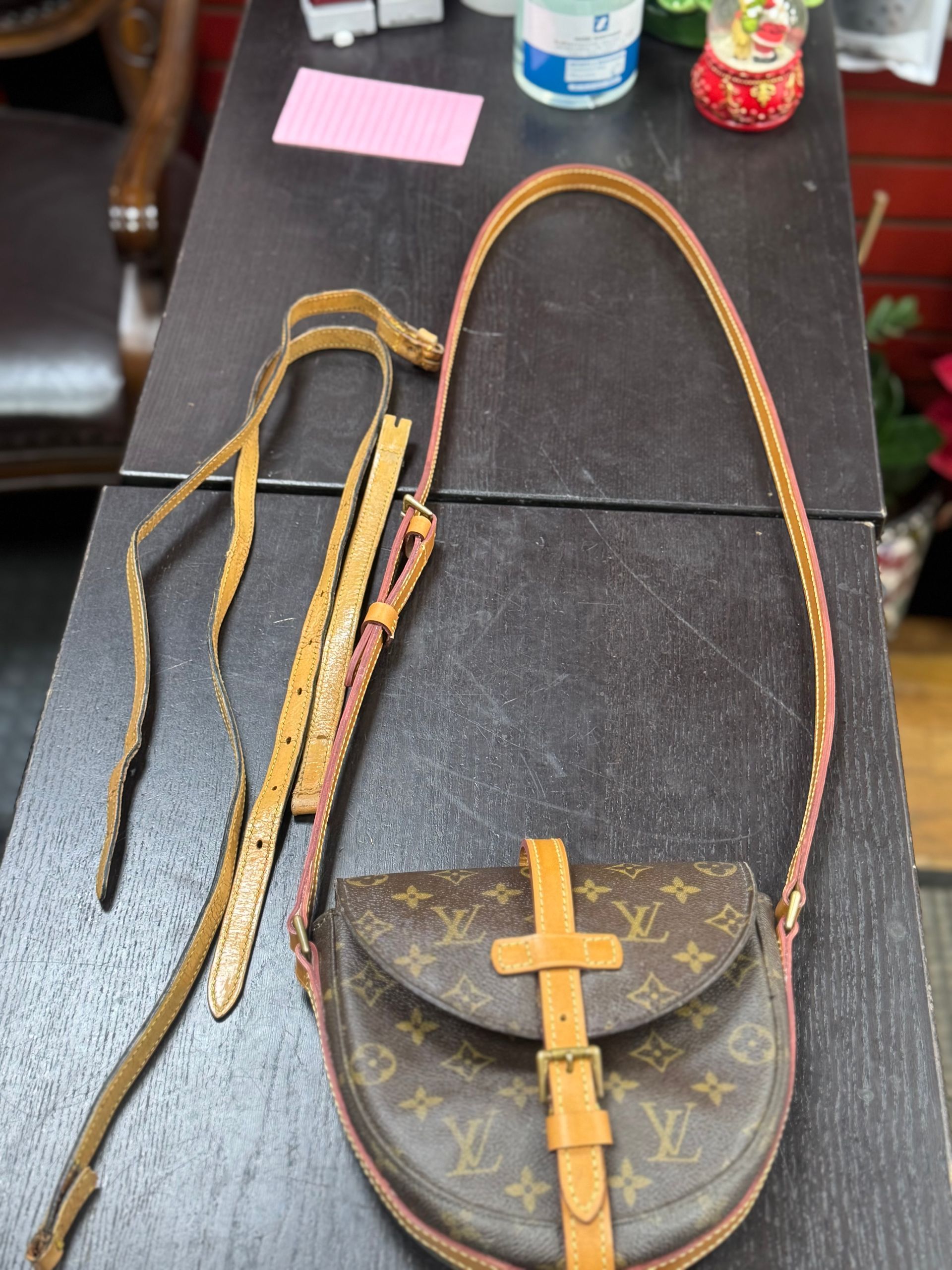 A Louis Vuitton monogram print handbag with a tan leather strap resting on a dark surface next to a detached strap.