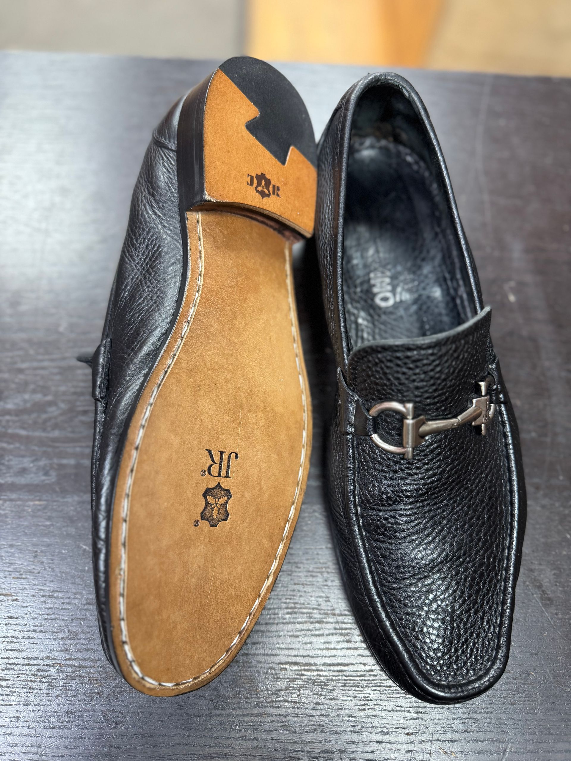 A pair of black leather loafers with silver bit hardware; one is shown sole-up, featuring a JR-branded leather bottom.