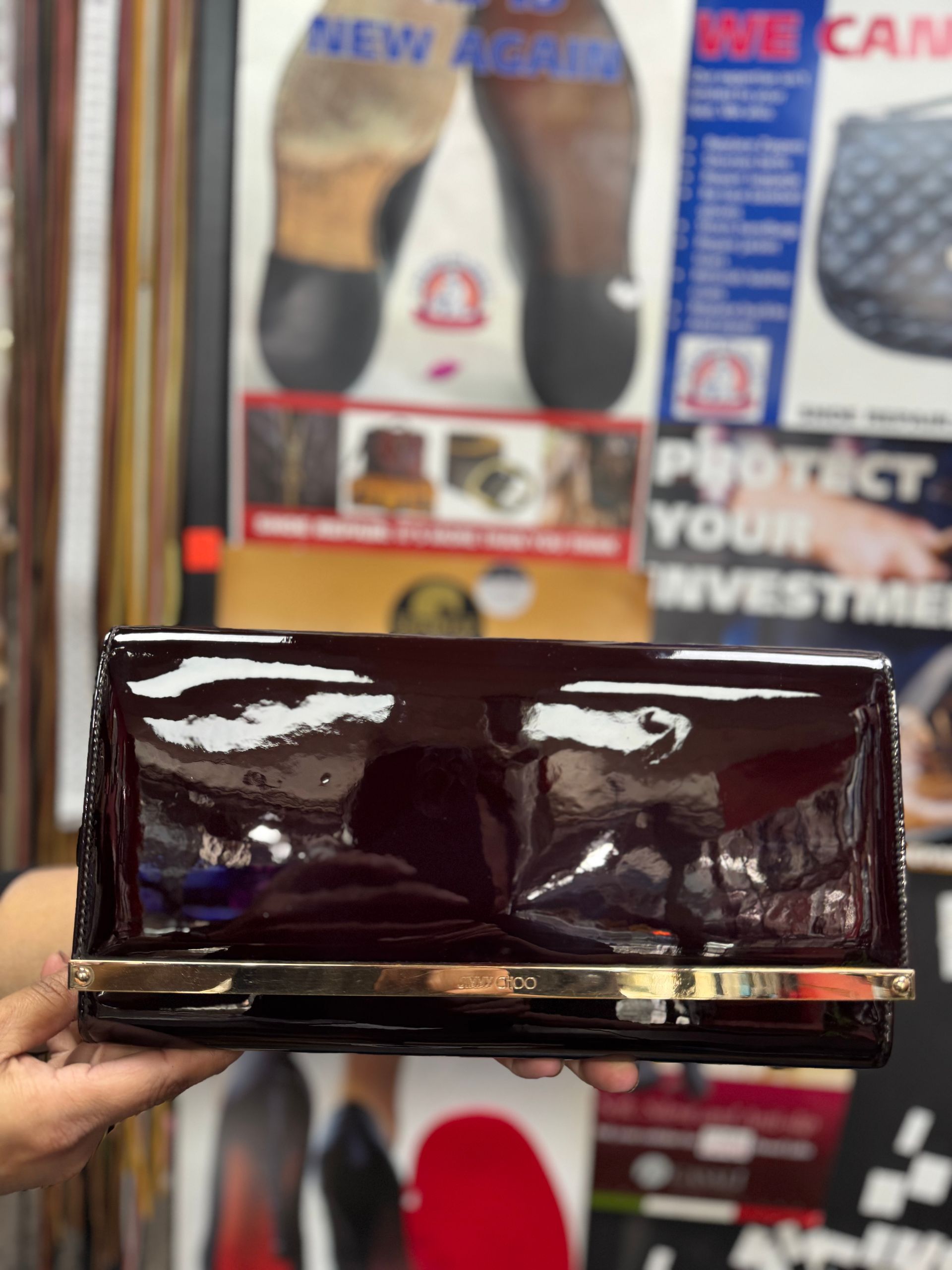 A person holds a shiny, dark burgundy patent leather clutch purse with a gold metal bar detail in front of a poster.