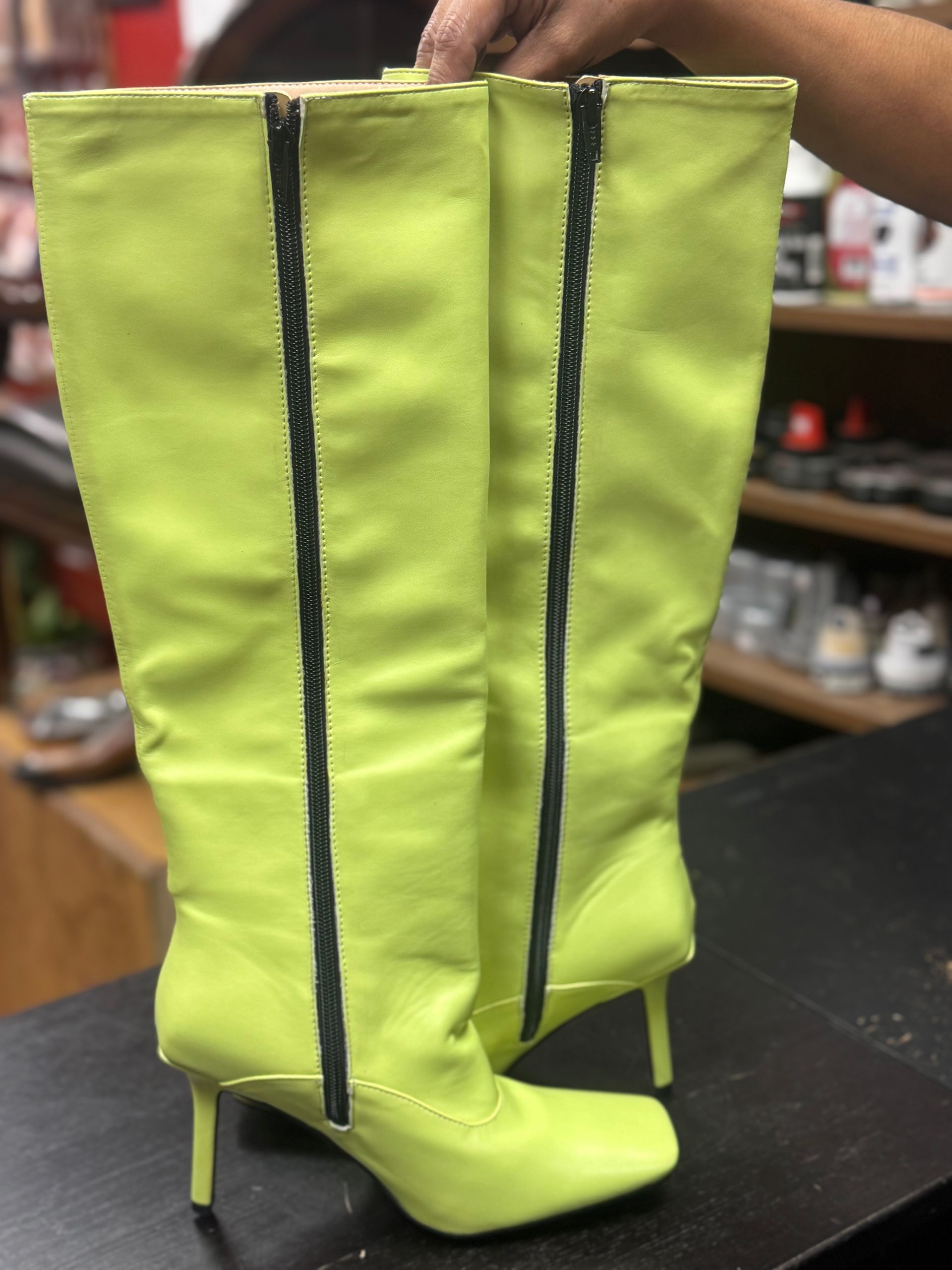 A pair of neon yellow, knee-high boots with thin heels and front zippers, held up against a blurry store background.