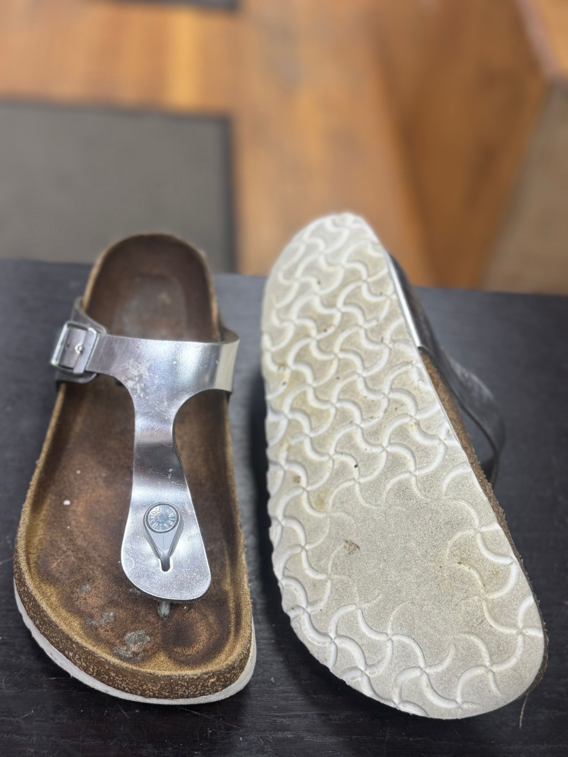 A pair of silver thong-style sandals resting on a dark surface, showing the footbed and the patterned white rubber sole.