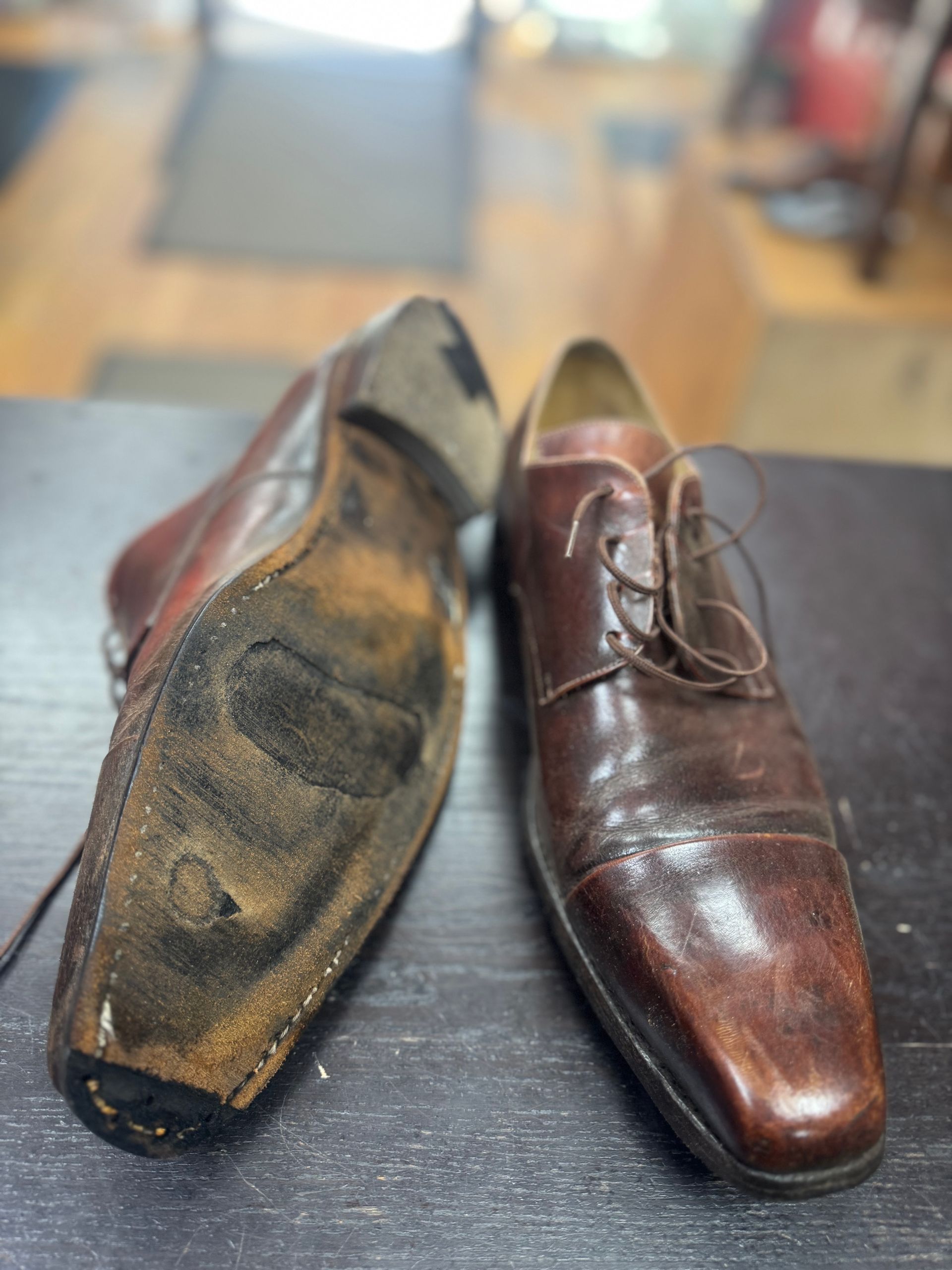 A pair of brown leather dress shoes on a dark surface, one placed sole-up to show significant wear and a hole.