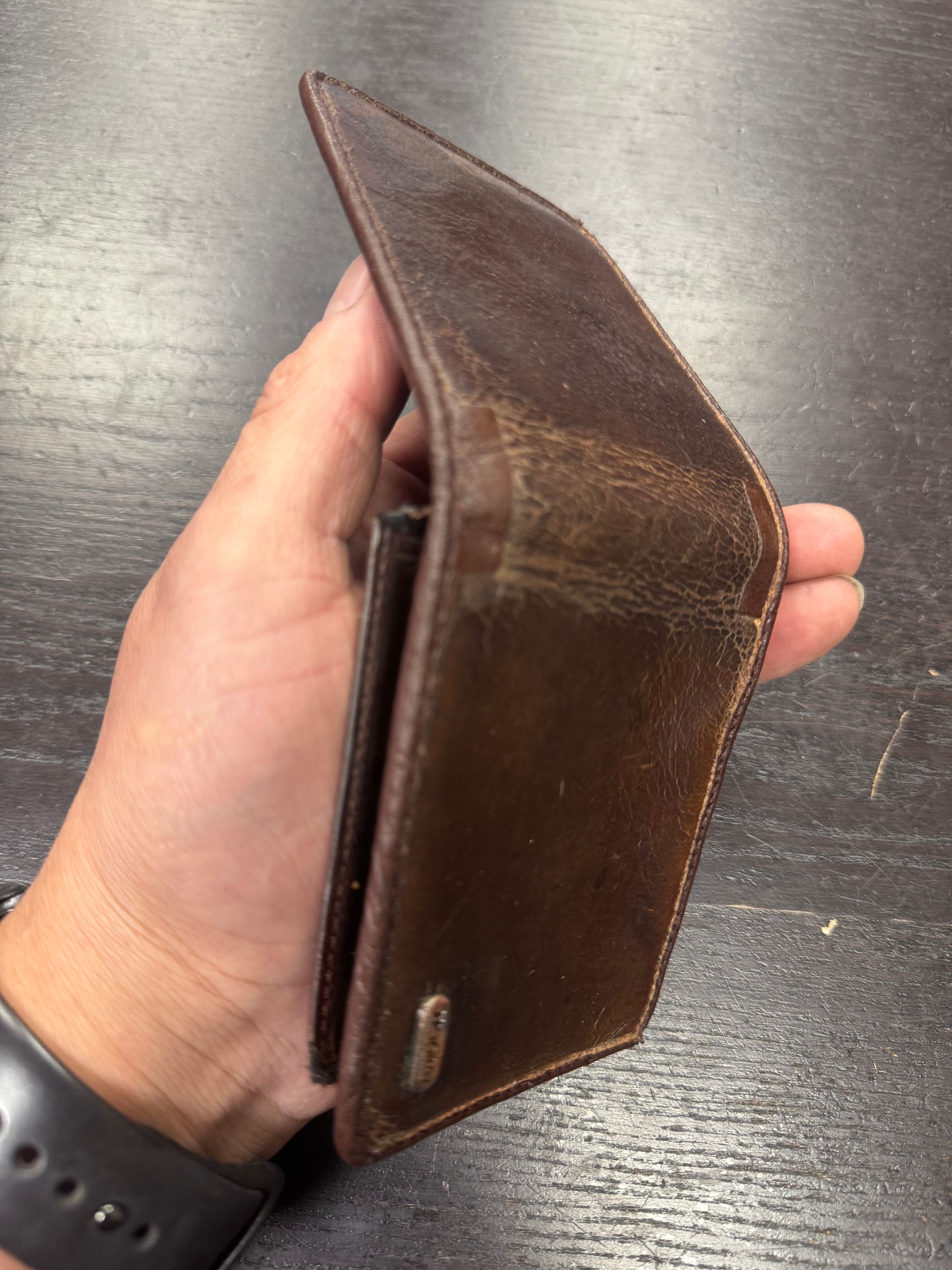 A hand holds a worn, dark brown leather bifold wallet against a dark wooden surface.