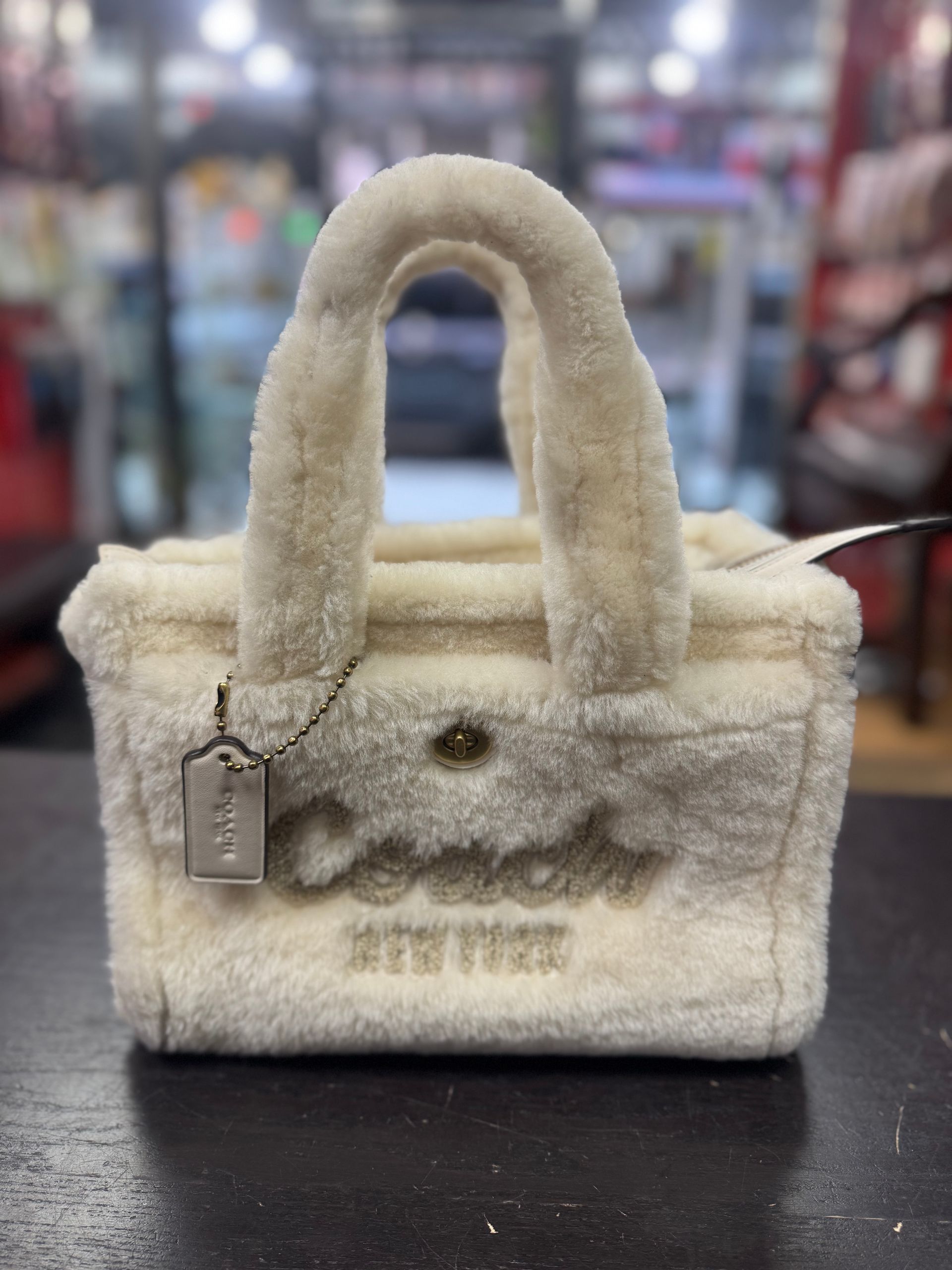 A cream-colored, faux-shearling Coach tote bag with embroidered logo lettering, sitting on a dark surface.