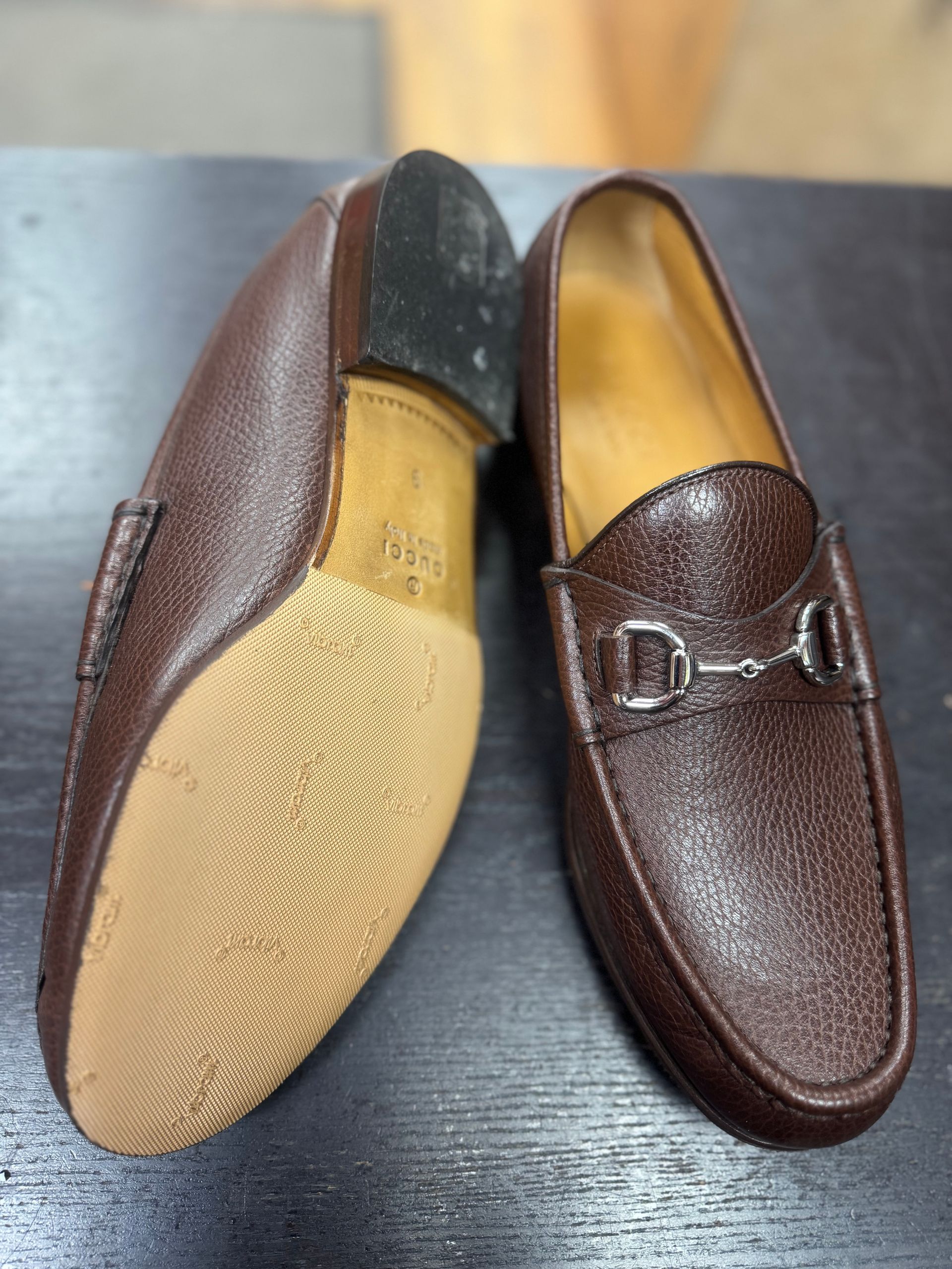 A pair of brown leather Gucci loafers with silver horsebit hardware, shown with one sole face-up on a dark surface.