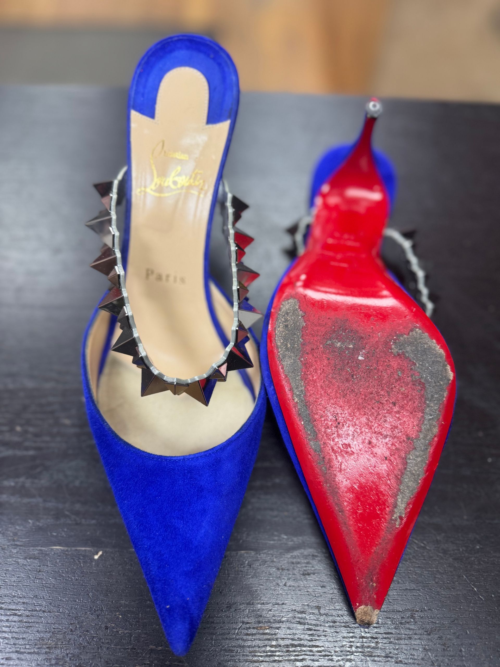 A pair of blue suede, spike-studded Christian Louboutin heels, one showing the interior and one showing the red sole.
