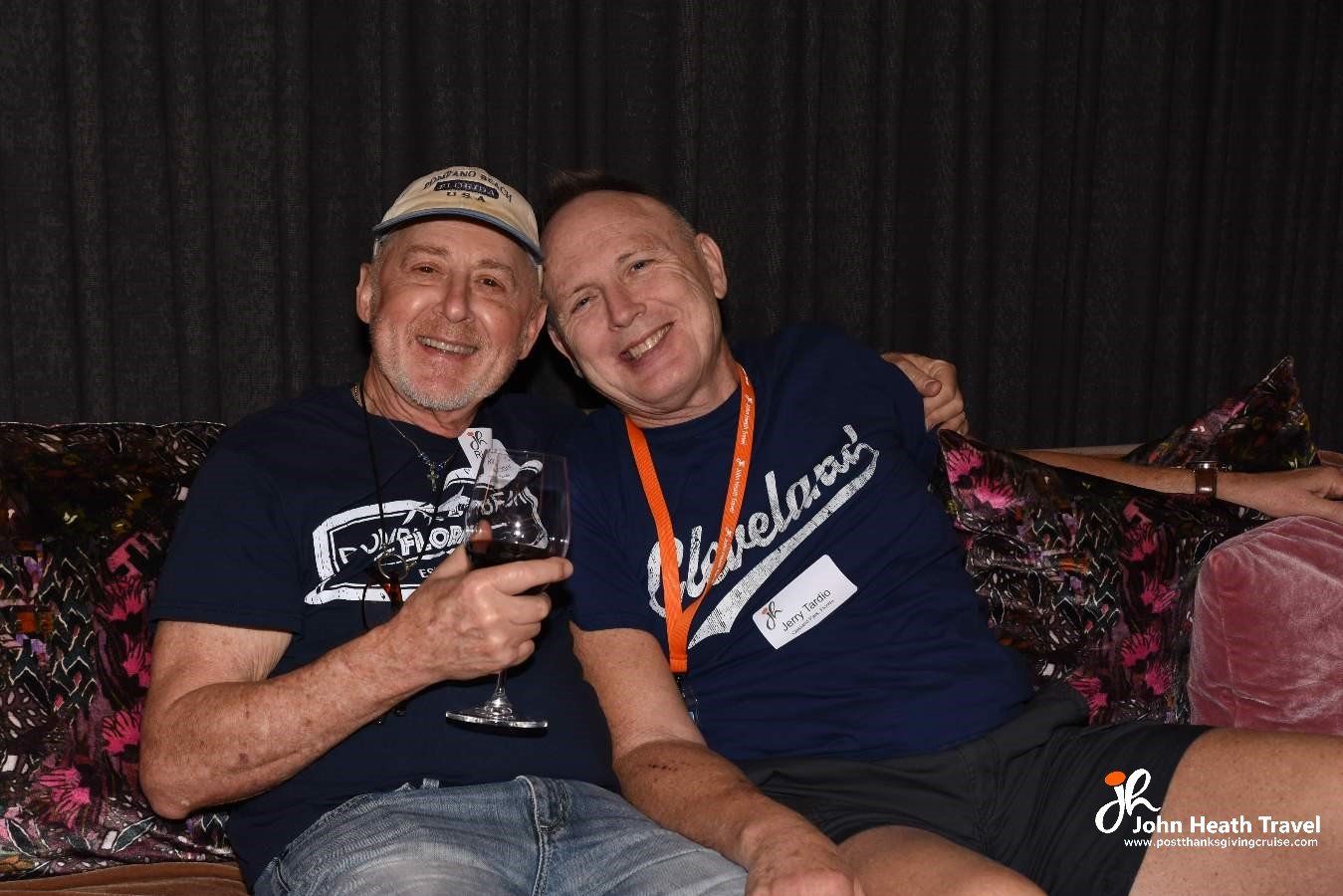 Two men are sitting next to each other on a couch and one of them is holding a glass of wine.