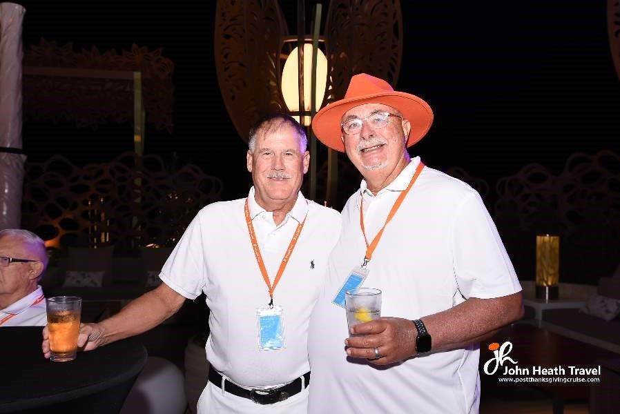 Two men are posing for a picture while holding drinks . one of the men is wearing an orange hat.