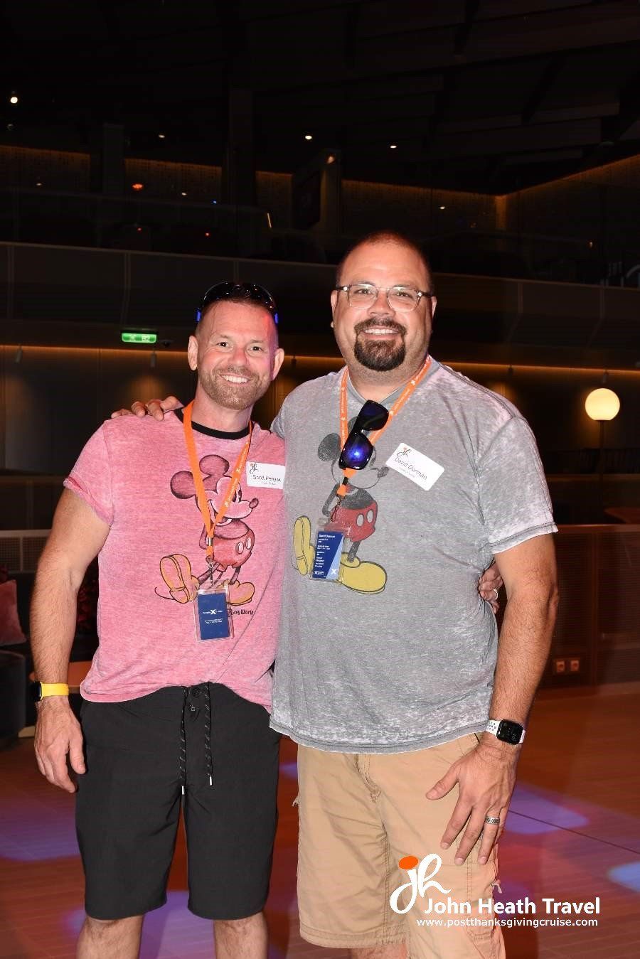 Two men are posing for a picture together in a room . one of the men is wearing a mickey mouse shirt.