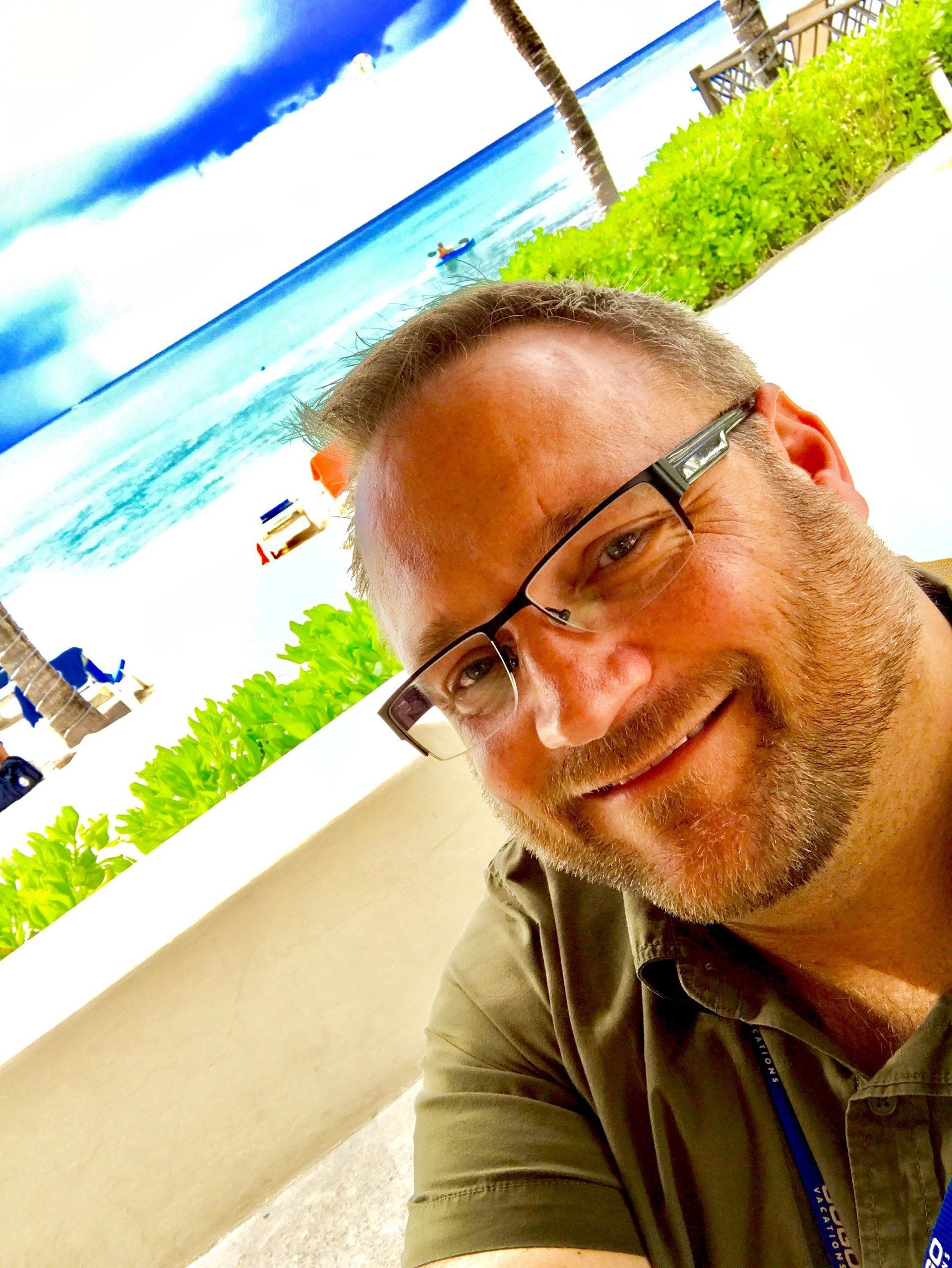 A man wearing glasses is smiling in front of a beach