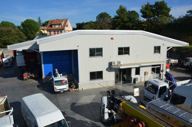 Un edificio industriale bianco con una porta di carico blu, circondato da furgoni e macchinari; esterno diurno.