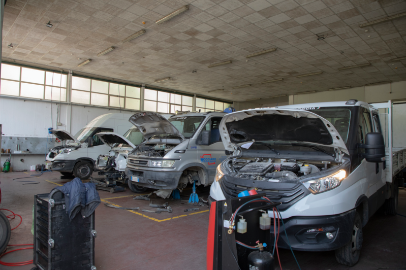 Veicoli in un'officina con cofani aperti, attrezzi e un tecnico.