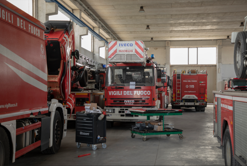 Camion dei pompieri parcheggiati all'interno di una caserma. Sono visibili veicoli, attrezzature e utensili rossi.