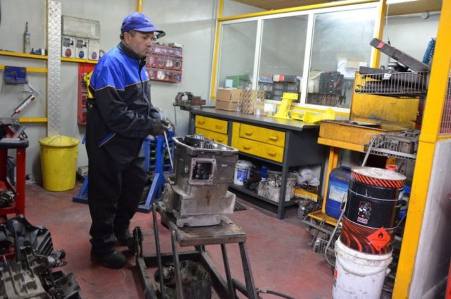 Un meccanico in tuta blu e nera tiene in mano un pezzo in un'officina. Sono visibili un banco da lavoro, attrezzi e contenitori.