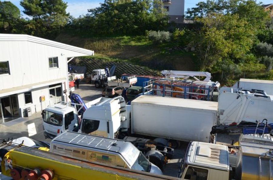 Veicoli parcheggiati presso una struttura, inclusi camion, furgoni e un'autocisterna. Una persona lavora su un veicolo.