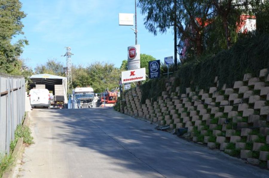 Una strada in pendenza con veicoli, un muro di contenimento in blocchi impilati e cartelli per un'attività commerciale.