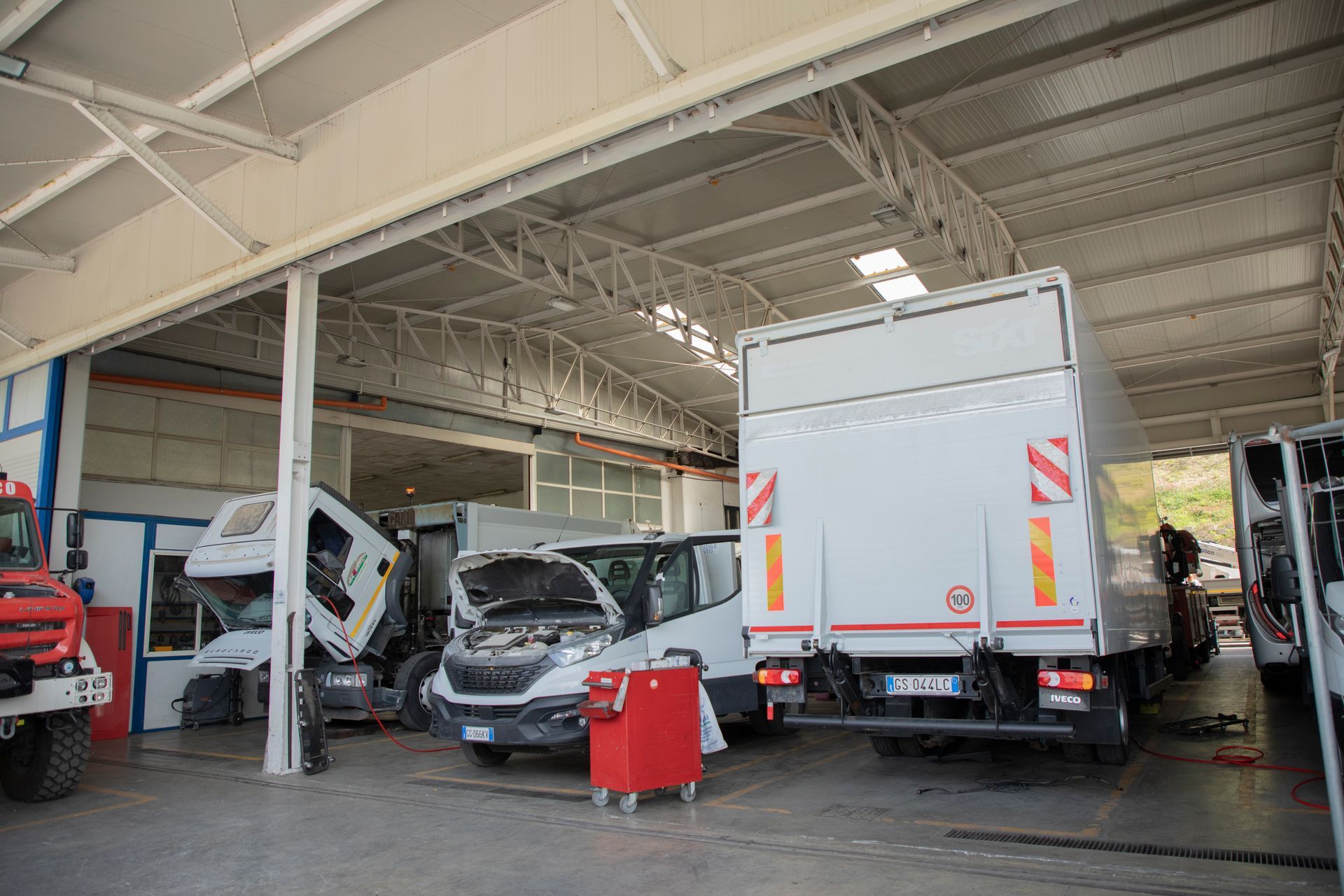 Camion e furgoni all'interno di un'officina con i cofani aperti e un carrello portautensili.