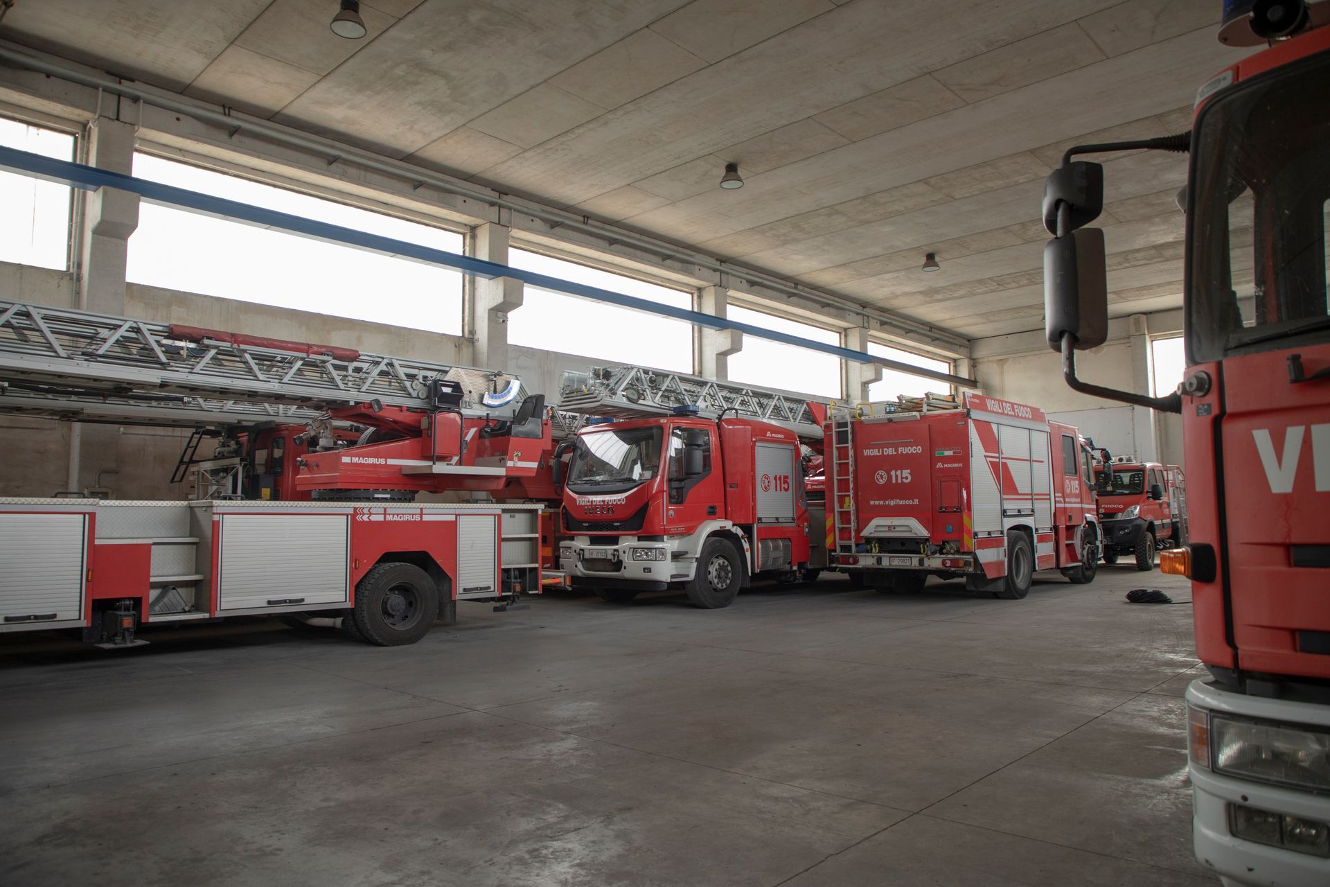 Camion dei pompieri parcheggiati all'interno di un ampio garage aperto. Sono visibili veicoli rossi e bianchi.