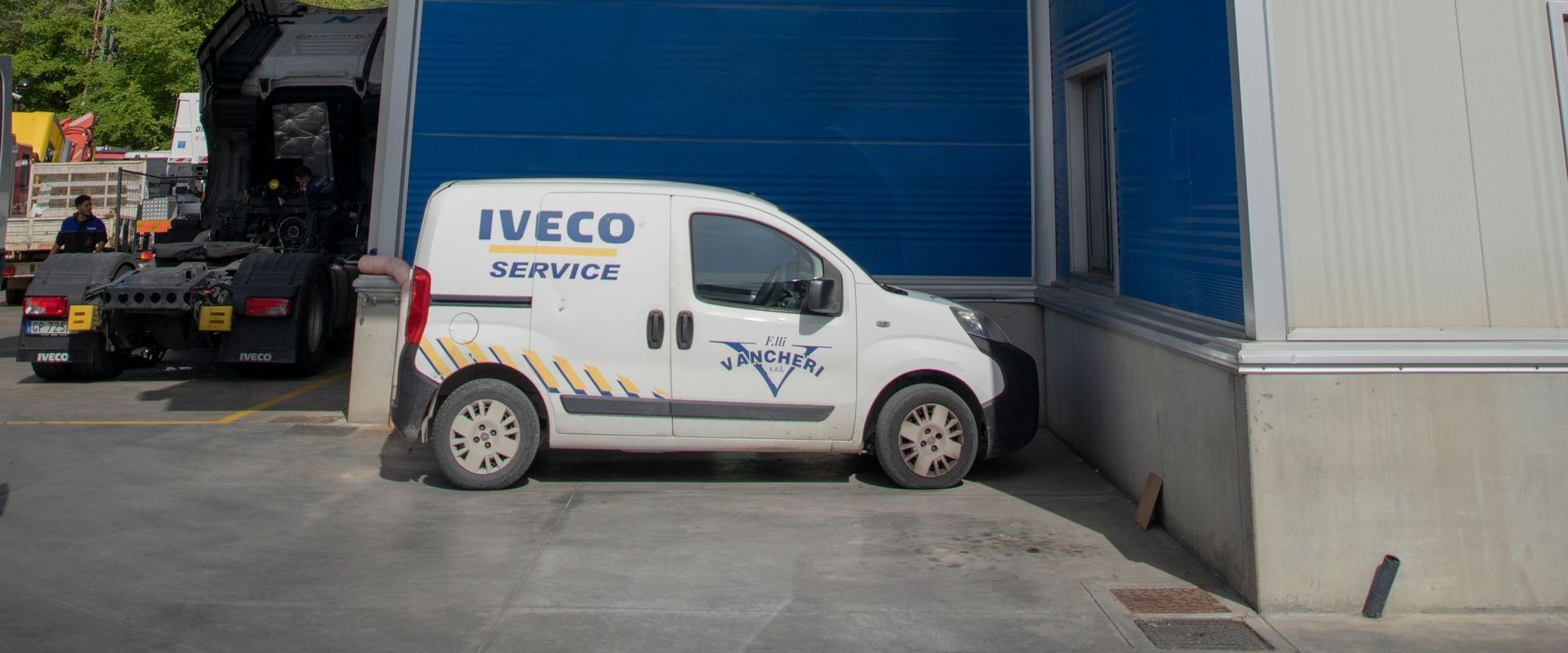 Un furgone Iveco bianco parcheggiato vicino alla porta blu di un magazzino, con un camion che fa retromarcia accanto.