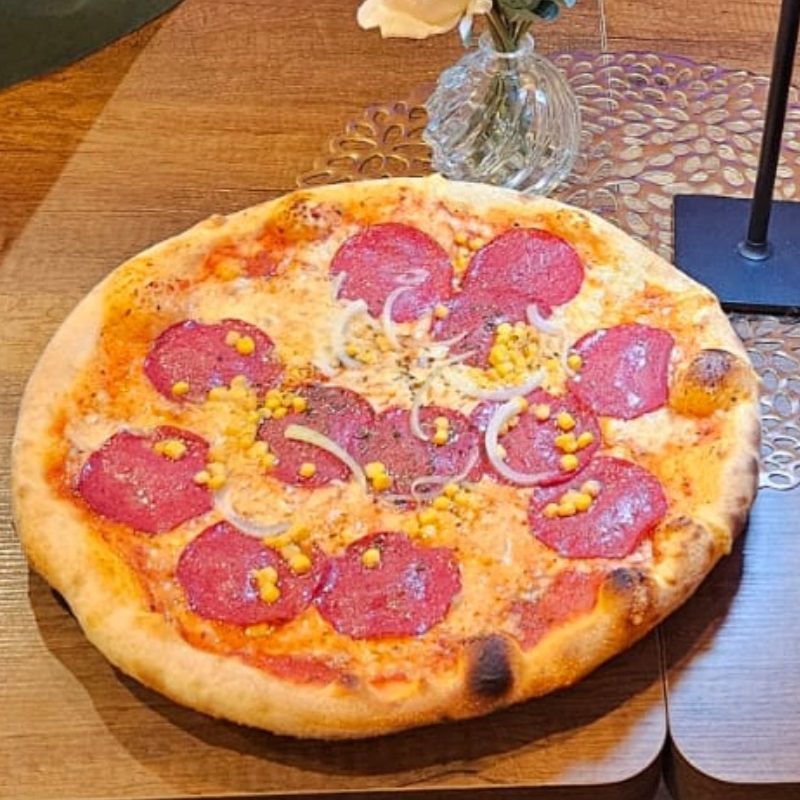 Peperoni-Pizza auf einem Holztisch mit Mais- und Zwiebelbelag neben einer Blumenvase.