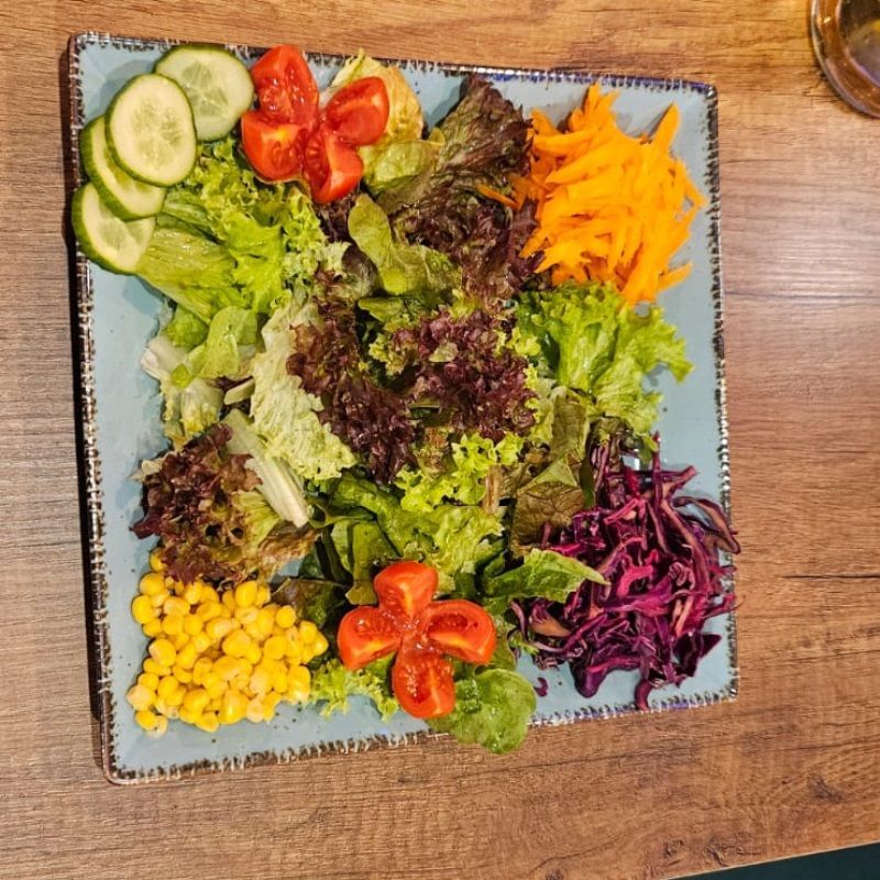 Bunter Salat mit Tomaten, Gurken, Karotten, Mais und Rotkohl auf einem blauen quadratischen Teller.