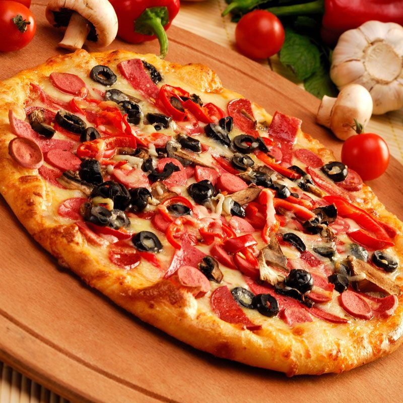 Pizza mit Wurst, Oliven und Paprika auf einem Holzbrett, umgeben von Zutaten.