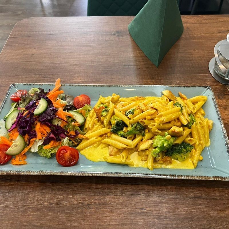 Nudelgericht mit Salat auf einem rechteckigen Teller. Die Nudeln sind mit einer gelben Sauce überzogen und mit Brokkoli garniert.